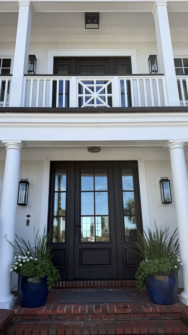 White two-story board-and-batten house with a black glass-pane front door, cobalt planters and lantern sconces